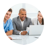 A man in blue scrubs, a man in a grey suit, and a woman in a white coat sit at a table and look at a laptop together